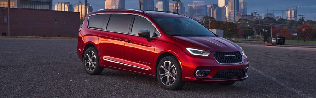 Red Chrysler Pacifica minivan parked in a lot with a city skyline in the background.