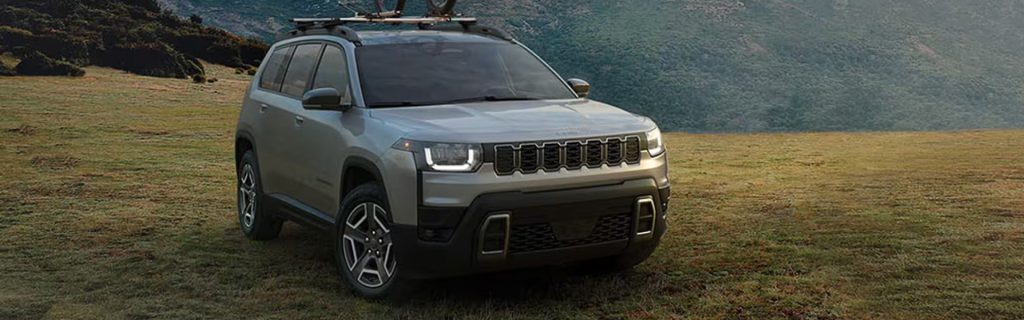 A silver Jeep SUV with a roof rack parked on a grassy field in front of hills.